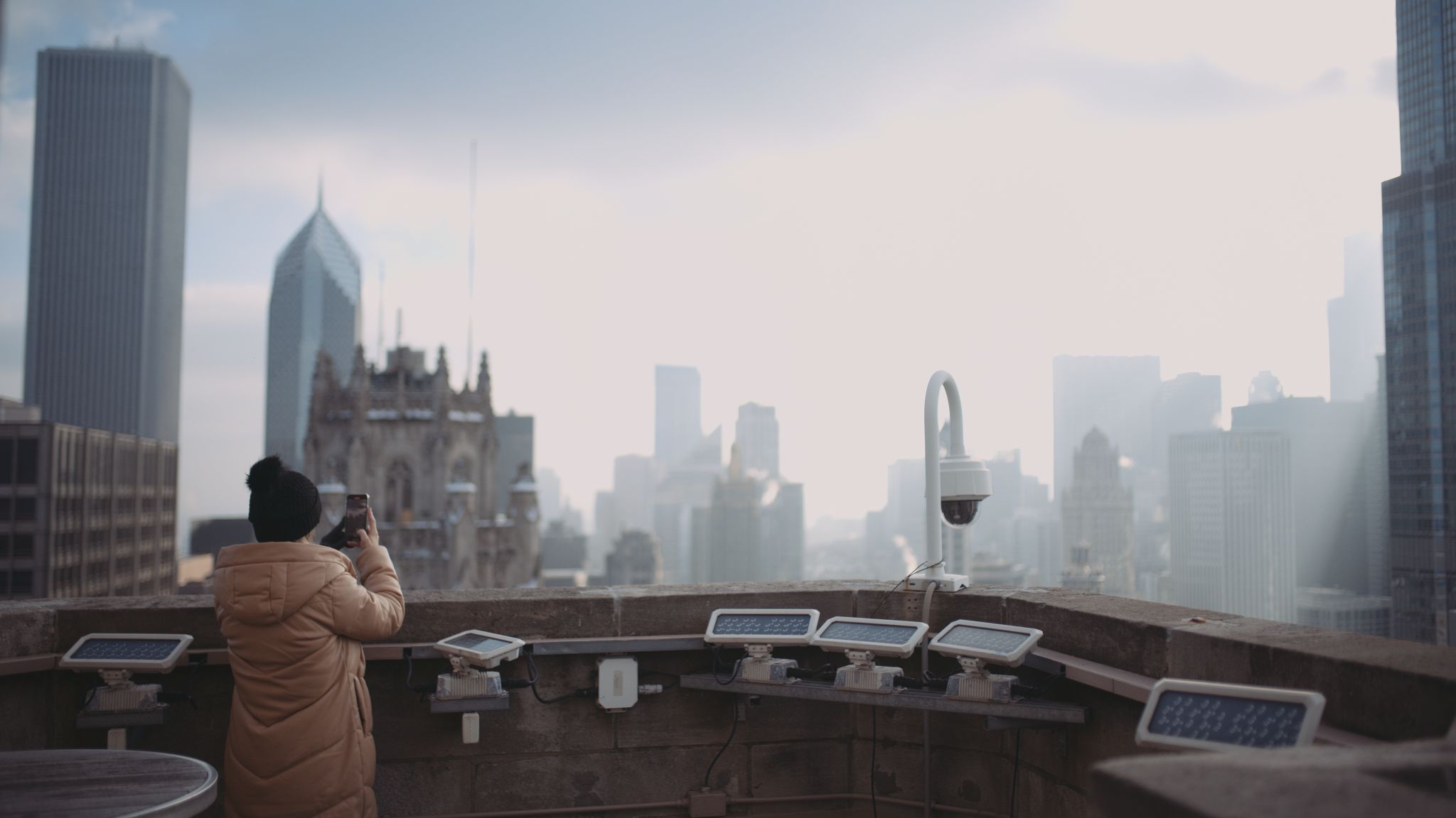 Woman in beige puffy jacket and black snow cap facing away from camera taking a picture of a skyline with her phone camera.