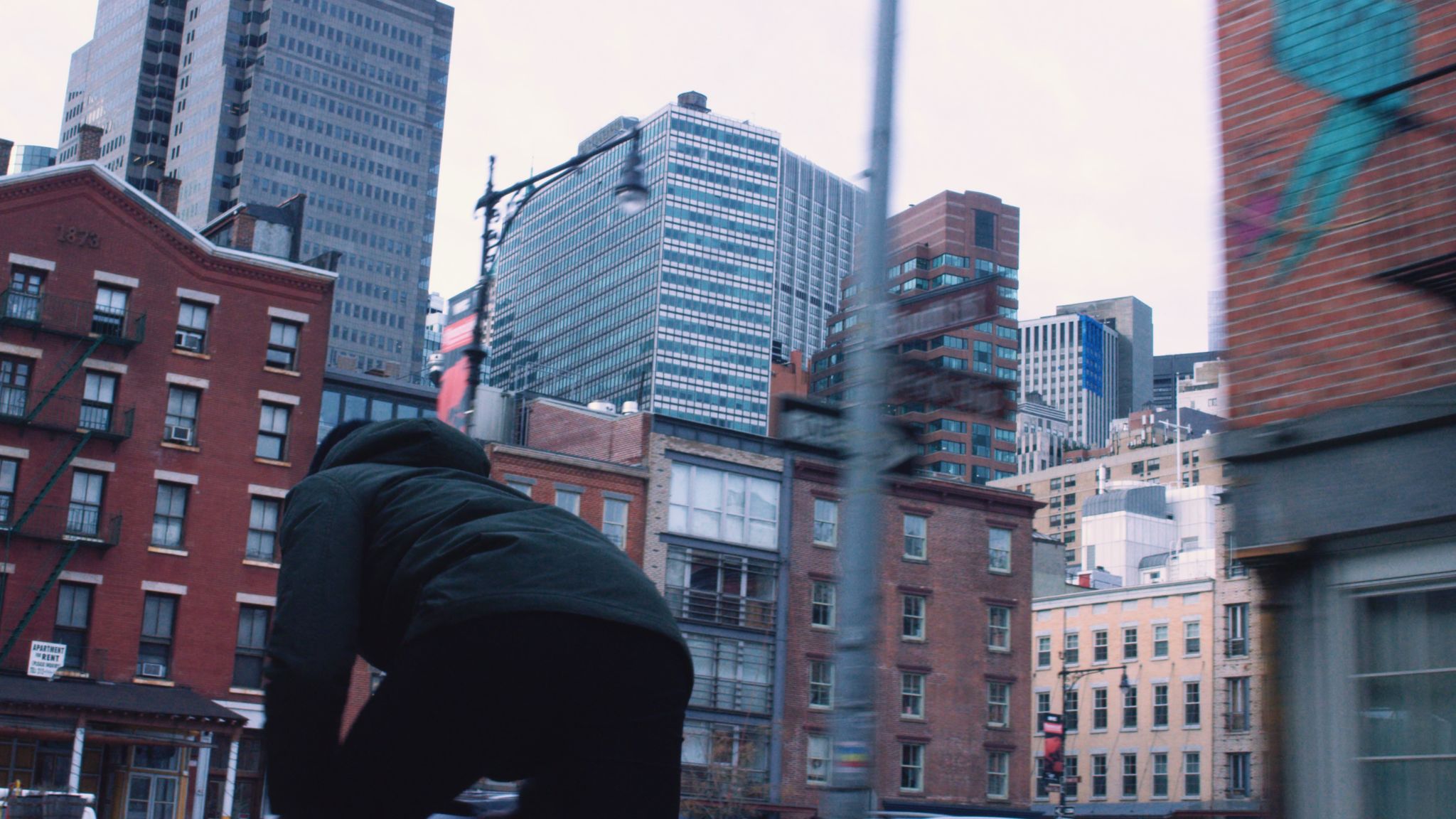 View from behind of man wearing olive green jacket and black pants biking through the city