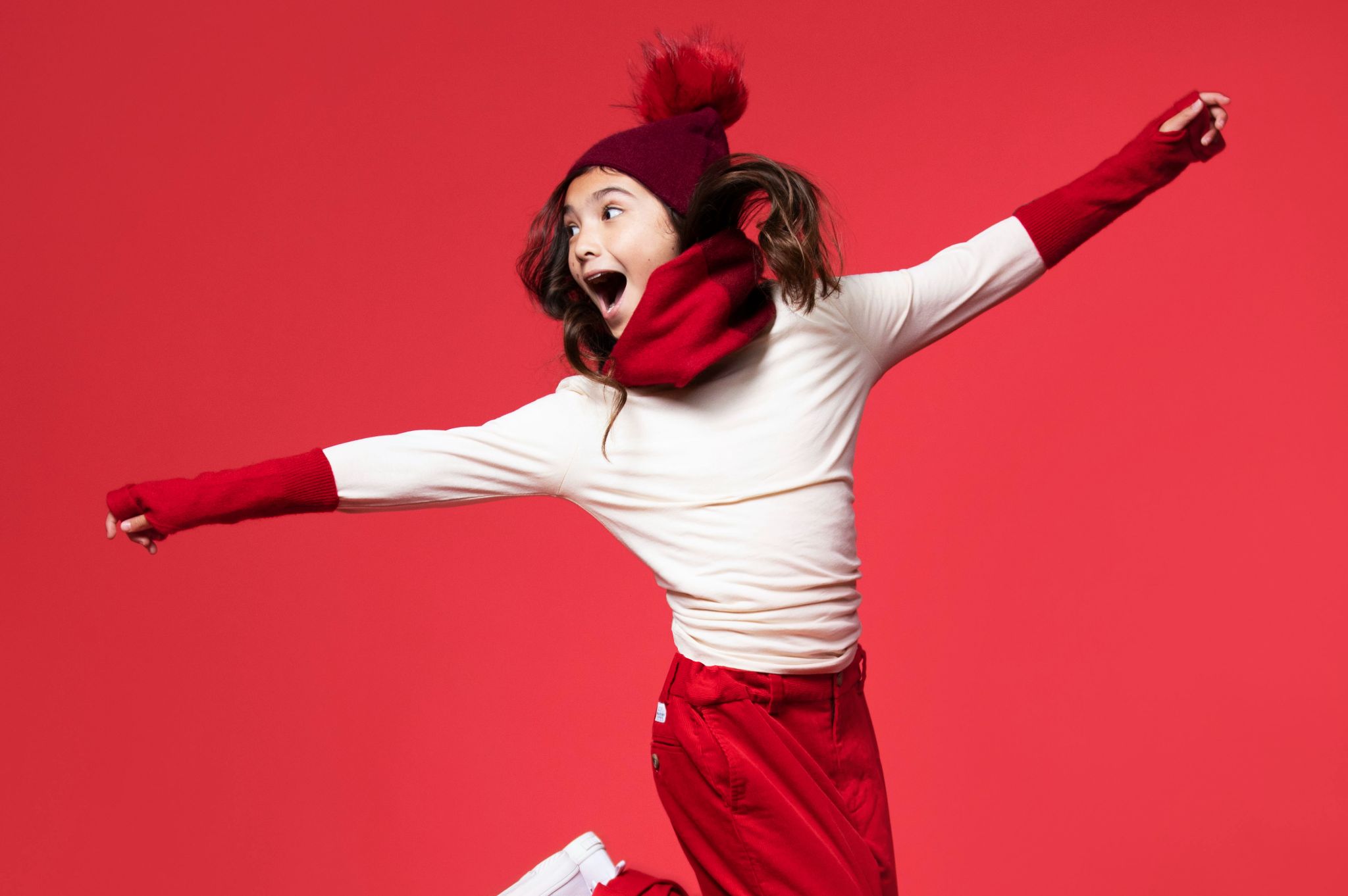 Why your Ecommerce Brand Should Start Creating TikTok Content Side profile of girl model wearing a red cap, scarf, hand sleeves and pants jumping and looking off to the side smiling against a red backdrop