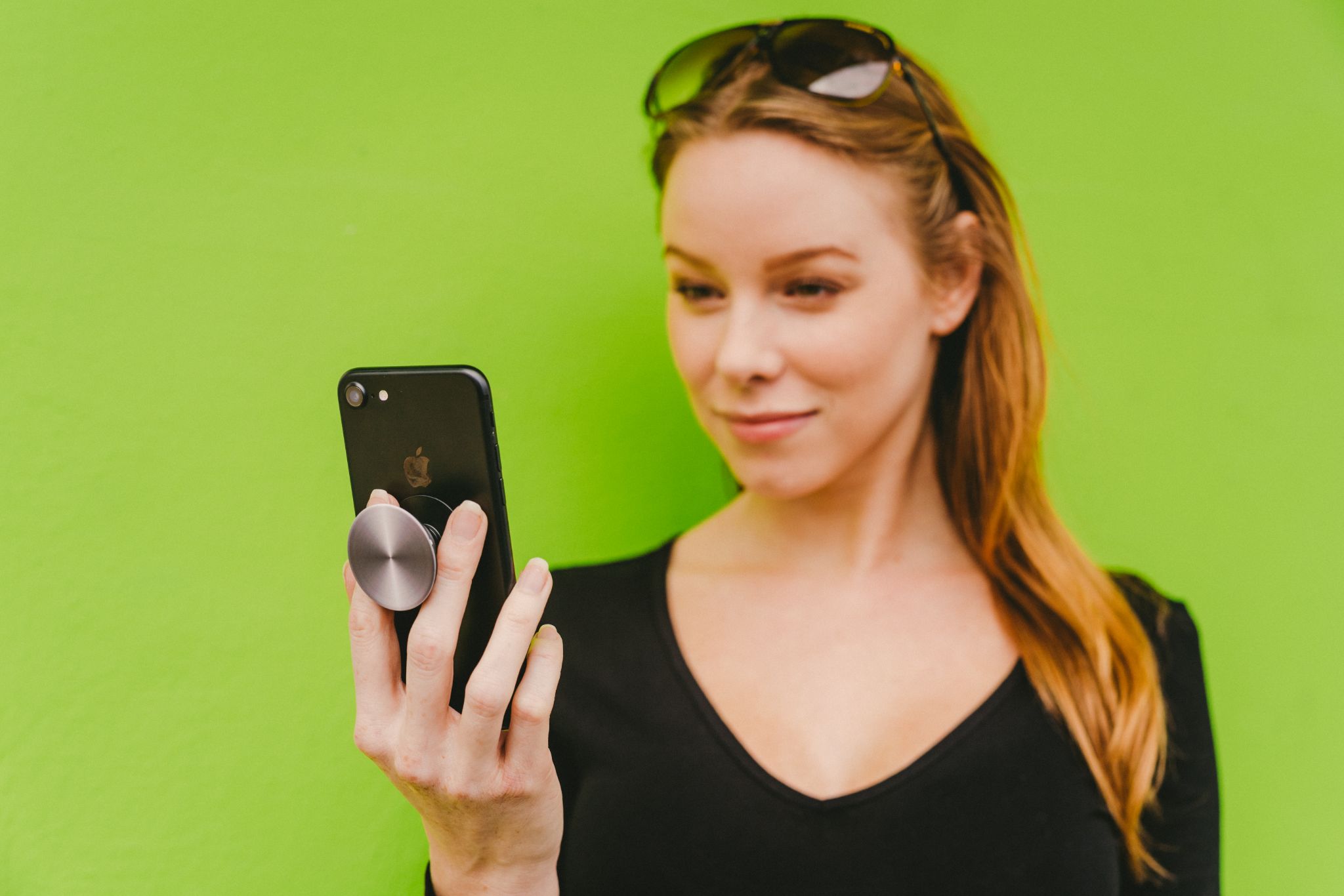 Closeup of person with long red hair wearing a black top using a cell phone leaning on a lime green colored wall