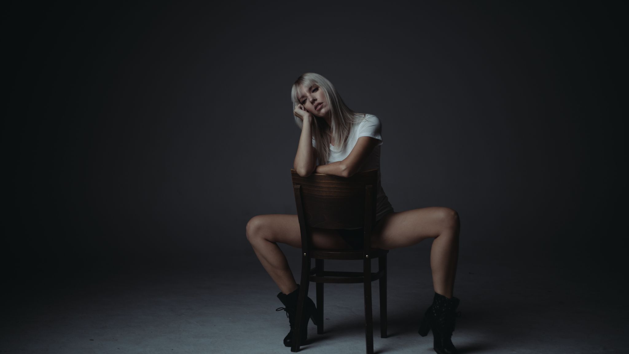 Female model with long, blond hair and white sleeveless shirt posing on a backwards chair facing camera.