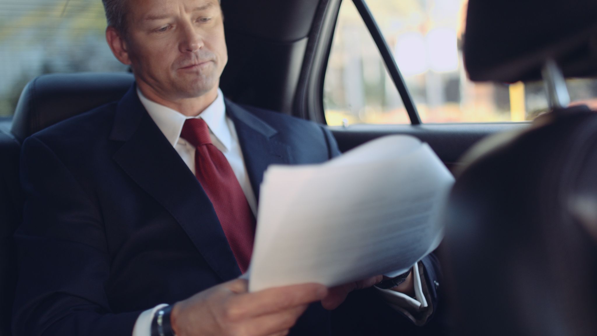 Closeup of man in a business suit with red tie sitting in the backseat of a car looking over papers