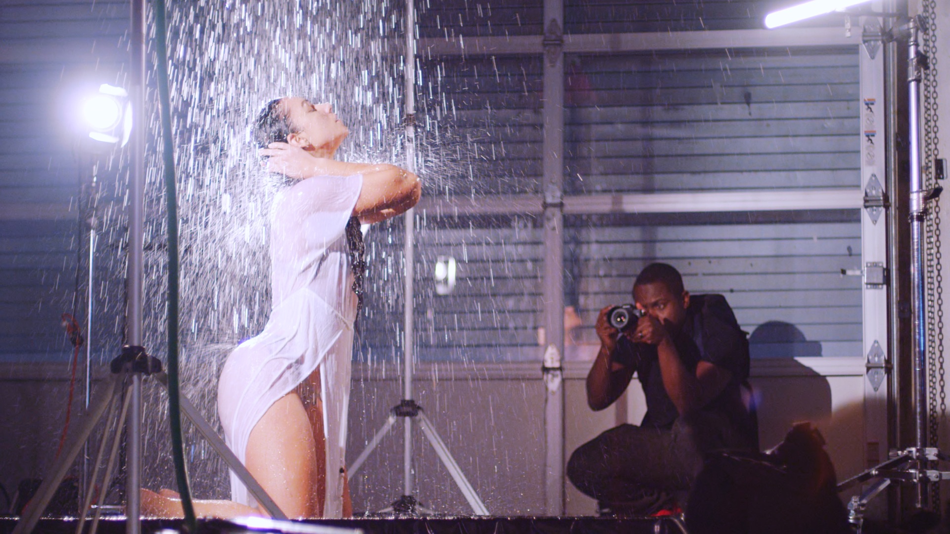 A female model clad in a see through outfit wearing jewelry under a shower on a set being photographed by an African American male.