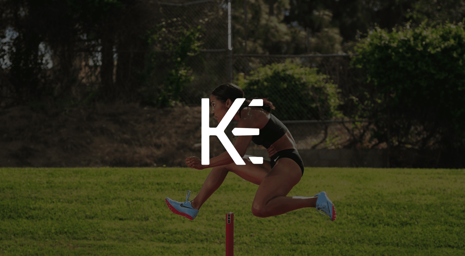 White KE logo against a dimmed background side view of a female track athlete jumping a hurdle