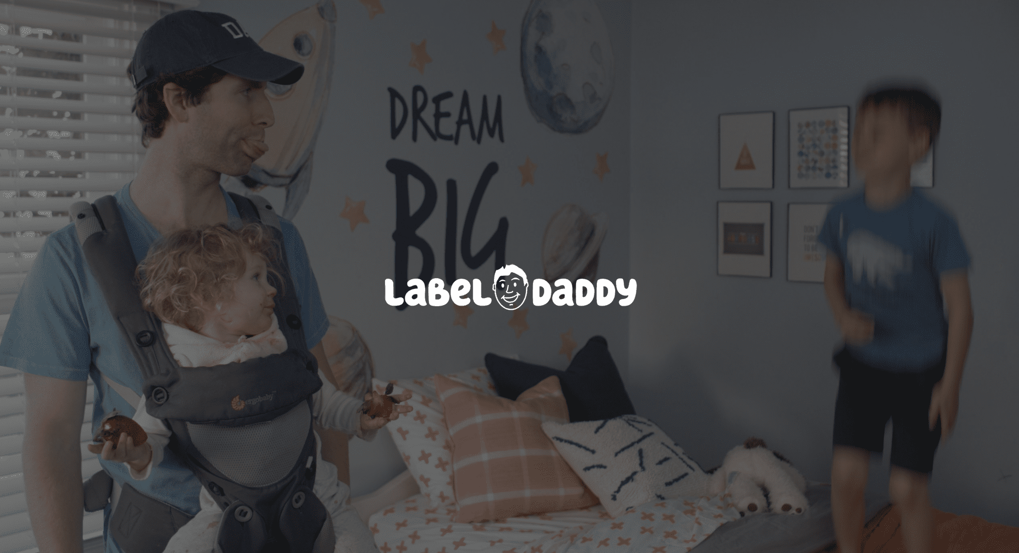 IU C&I Studios Page Label Daddy Featured Image White Label Daddy logo with background of Daddy doing a raspberry at boy standing on bed while carrying a girl in a chest carrier who is holding strawberries