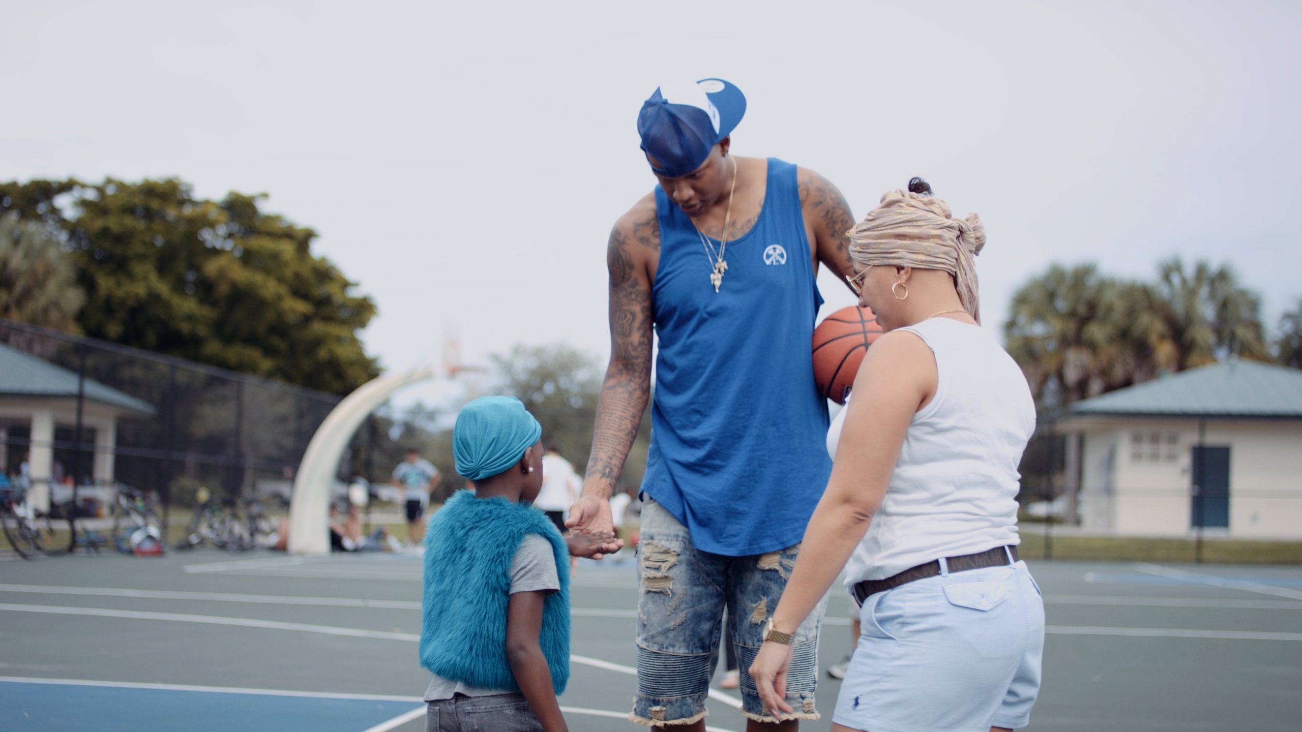 Charlie V wearing blue shirt, jean shorts and blue and white ball cap holding a basketball talking to girl wearing blue fur vest and jeans along with blue bandana while a woman looks on wearing white tank top and jean shorts