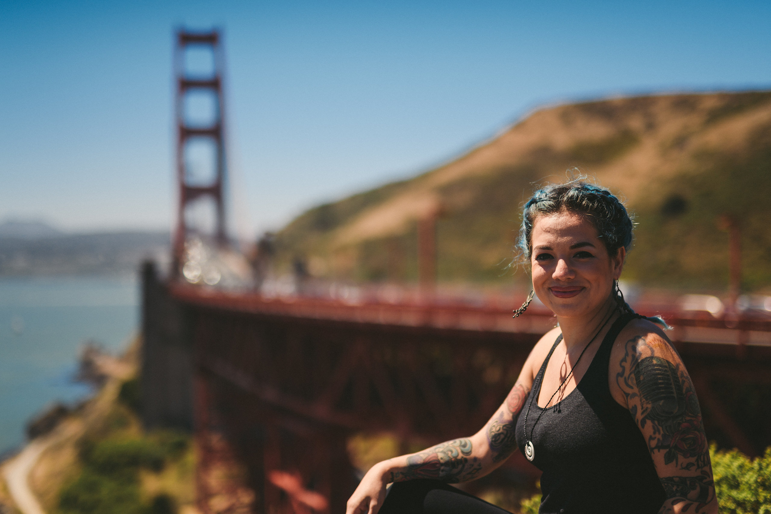 Tattooed woman with blue tint hair and wearing a black tank top smiling and posing by the San Francisco Bridge