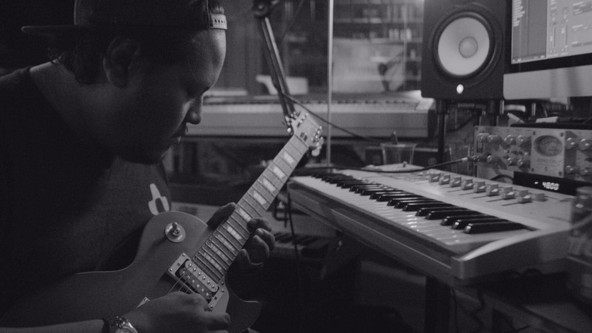 Black and white side profile of man playing a guitar by a keyboard in a studio