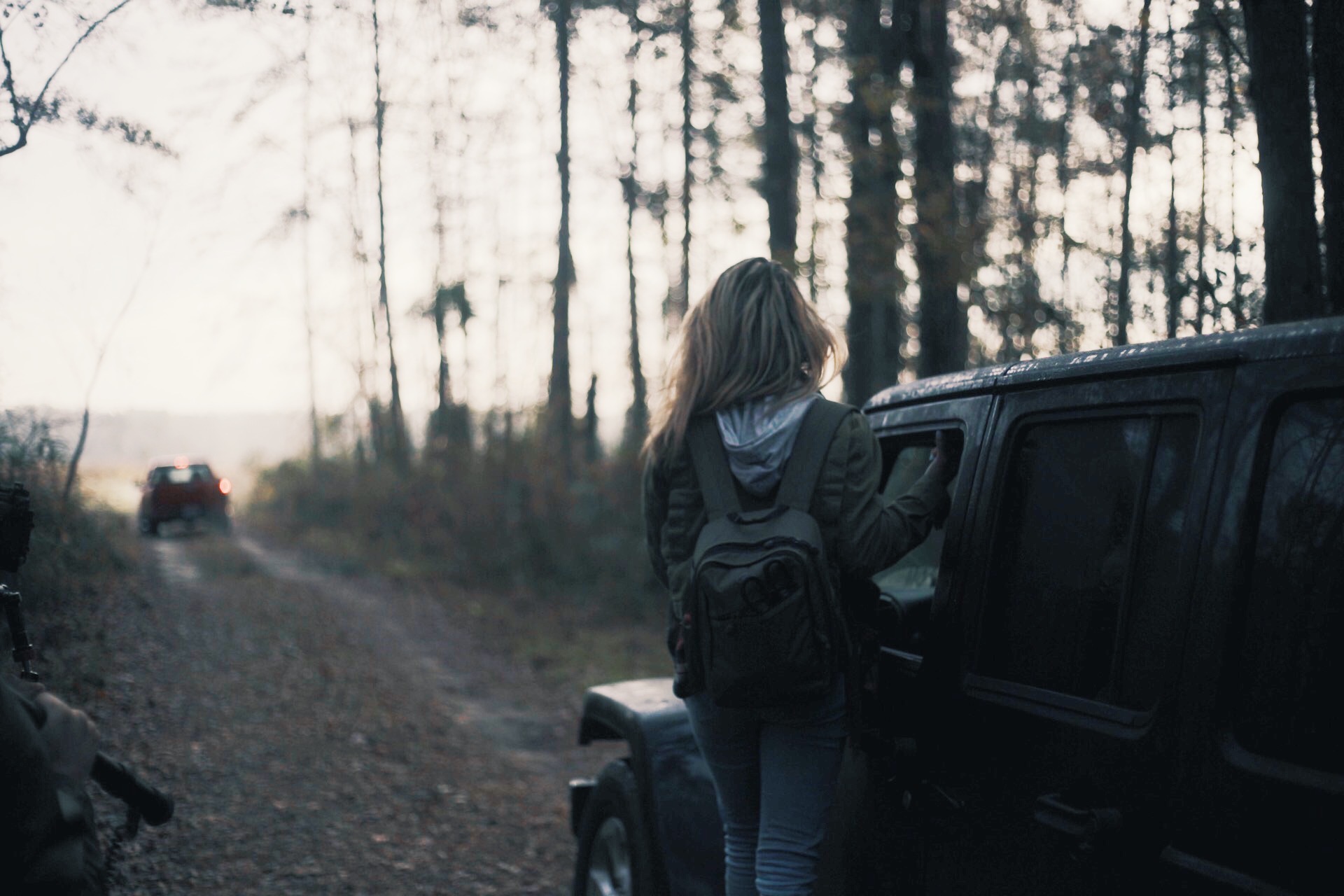 View from behind of woman with long blond hair wearing a backpack standing by a jeep looking at a red pickup truck up the road