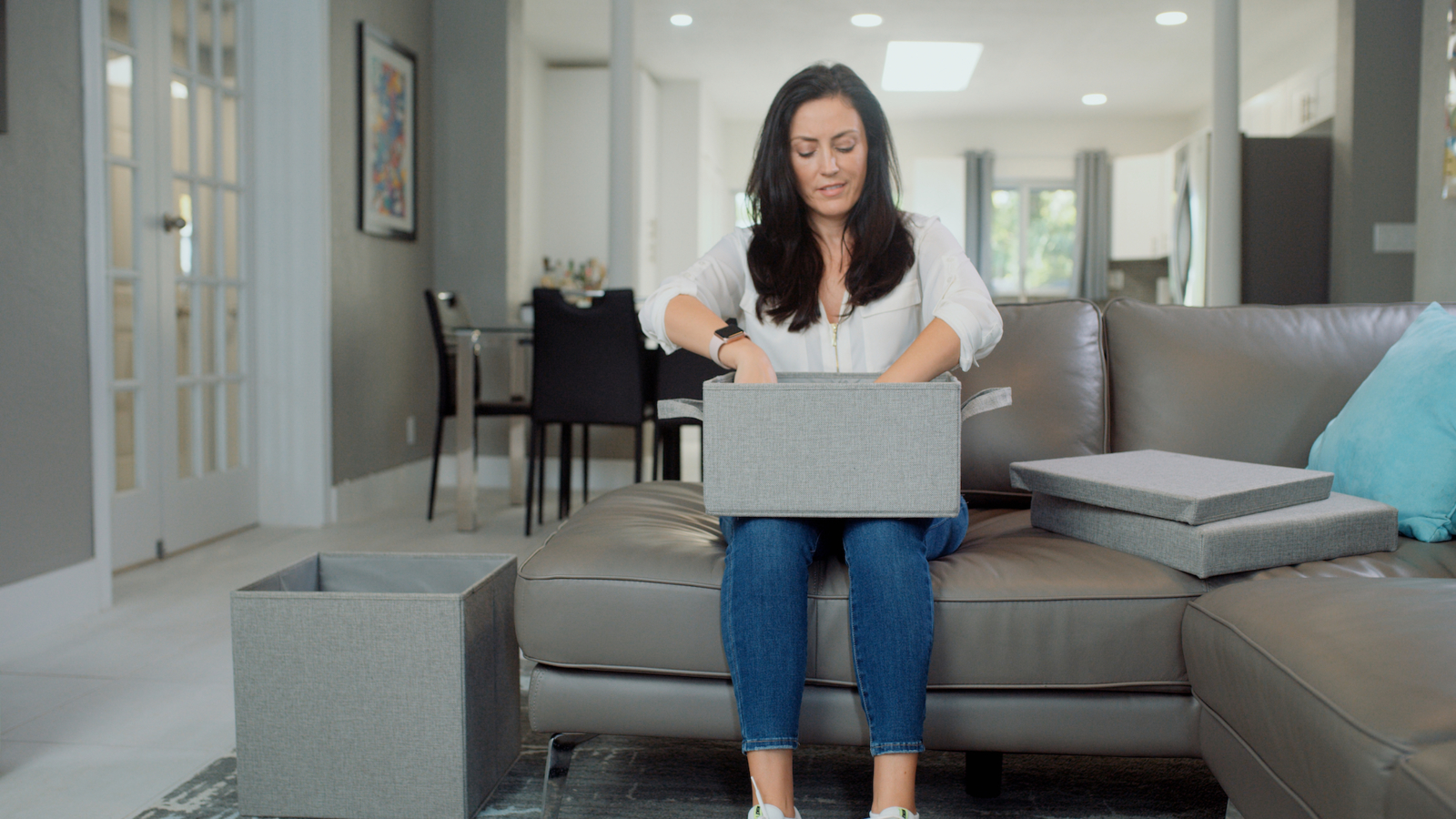 Woman with long black hair wearing a white top and blue jeans sitting on a gray leather couch going through a box