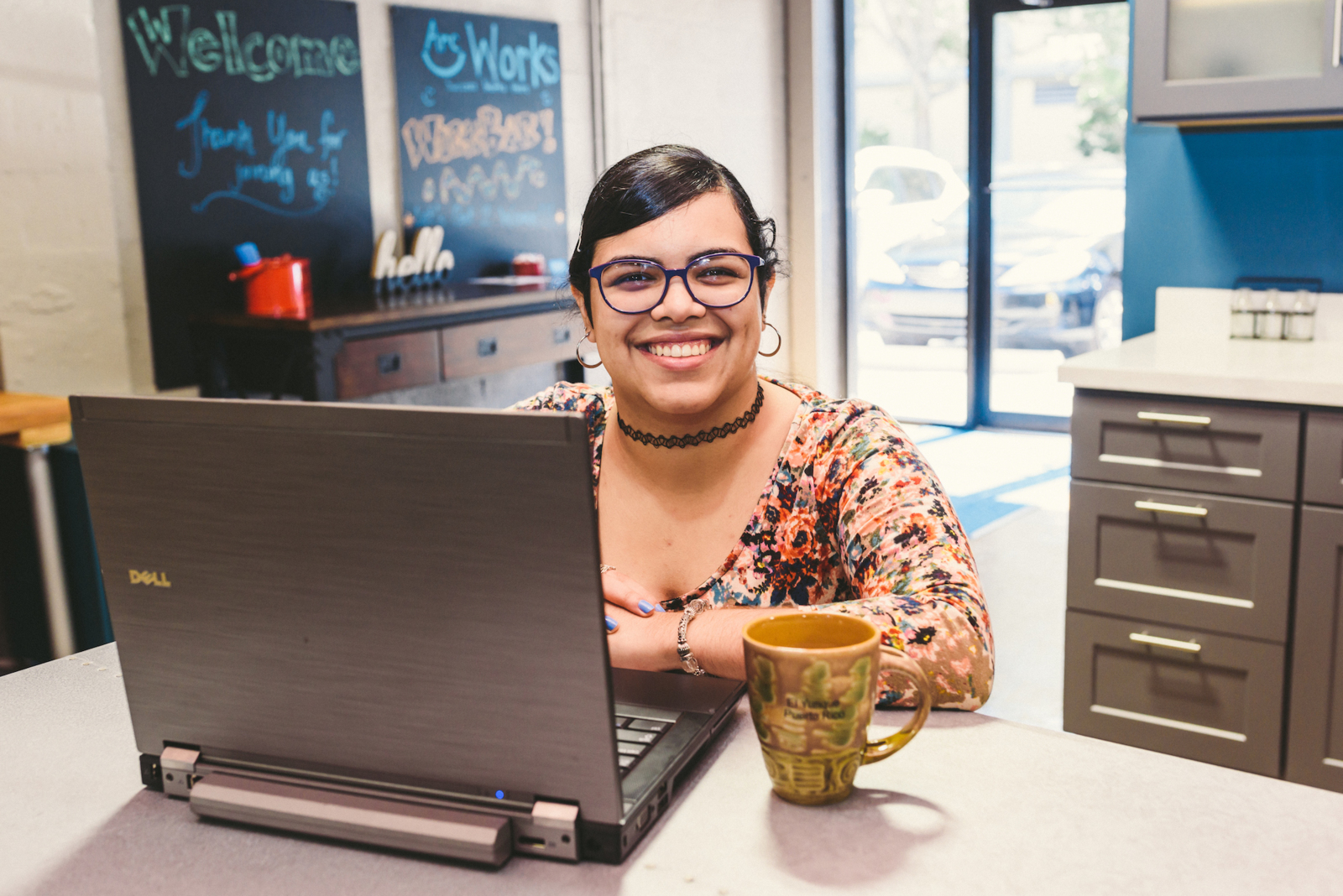 Woman using a laptop with a brown and green coffee cup next to it wearing a colorful top smiling for the camera
