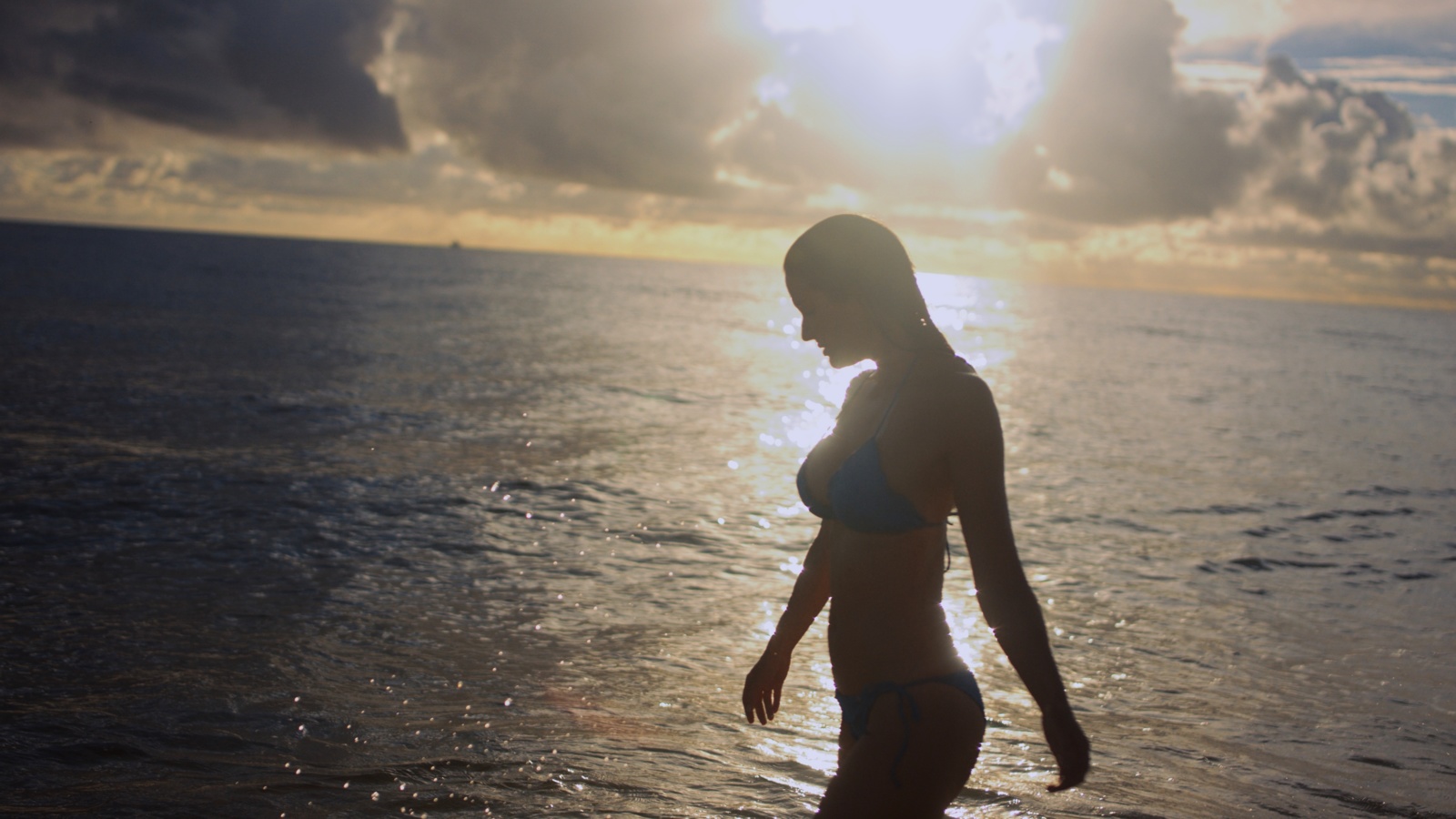 Side profile of woman wearing a bikini walking along a beach at sunset