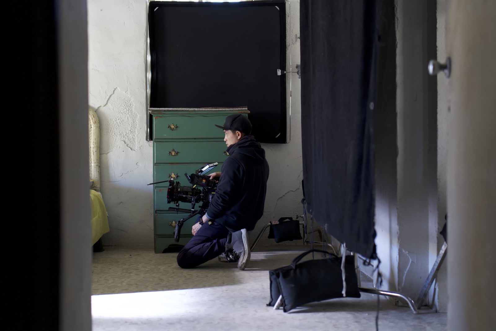 Side profile of a male crew member wearing a black cap using a video camera in a room with a bed