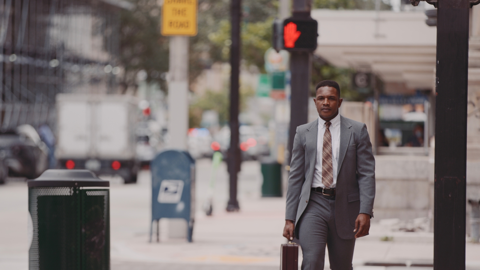 African American man in a gray suit with white shirt and brown tie walking down a city street carrying a brown briefcase