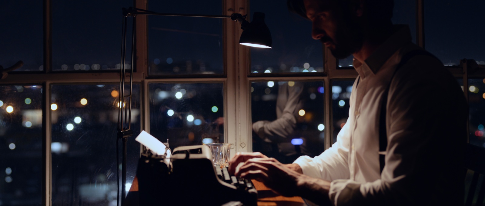 Side profile of a man wearing a white shirt and suspenders using a typewriter at night by windows overlooking a city
