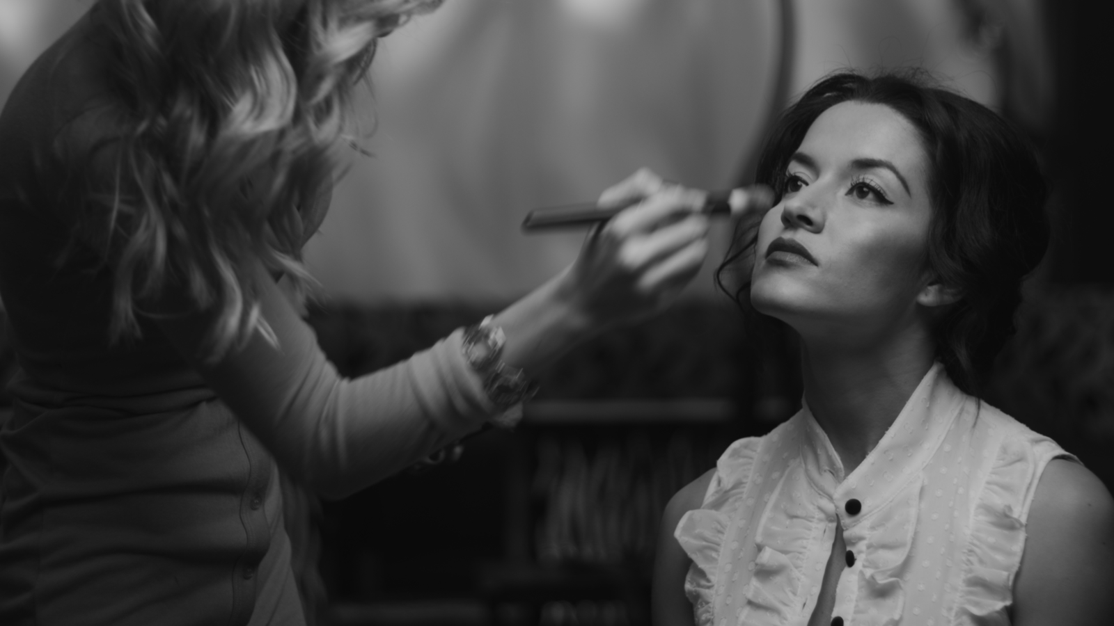 Black and white side profile of female makeup artist applying makeup to woman with long hair wearing a white dress top