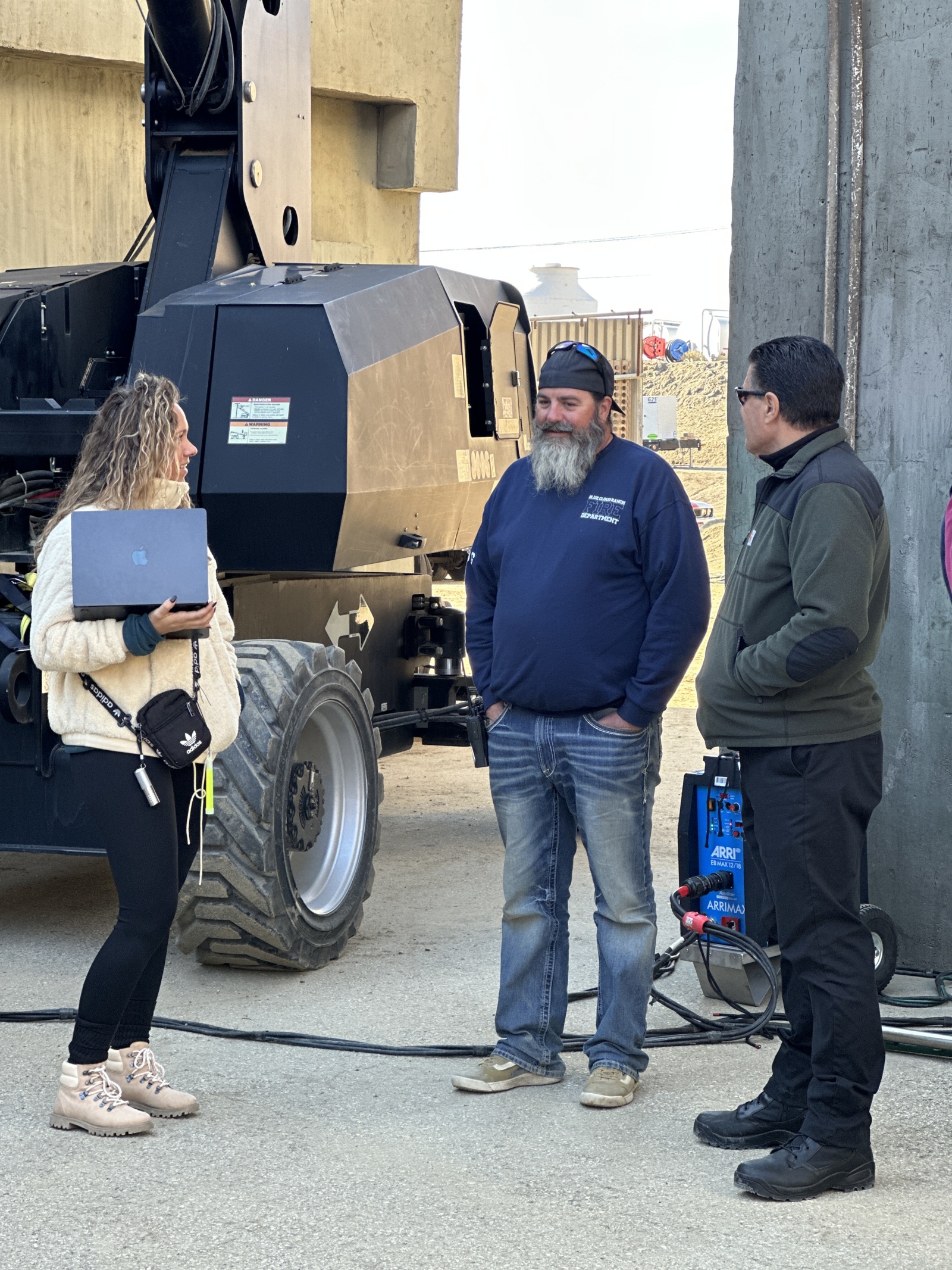 Man wearing black cap backwards, dark blue sweater, jeans and sneakers talking with man and woman holding a laptop on a construction site