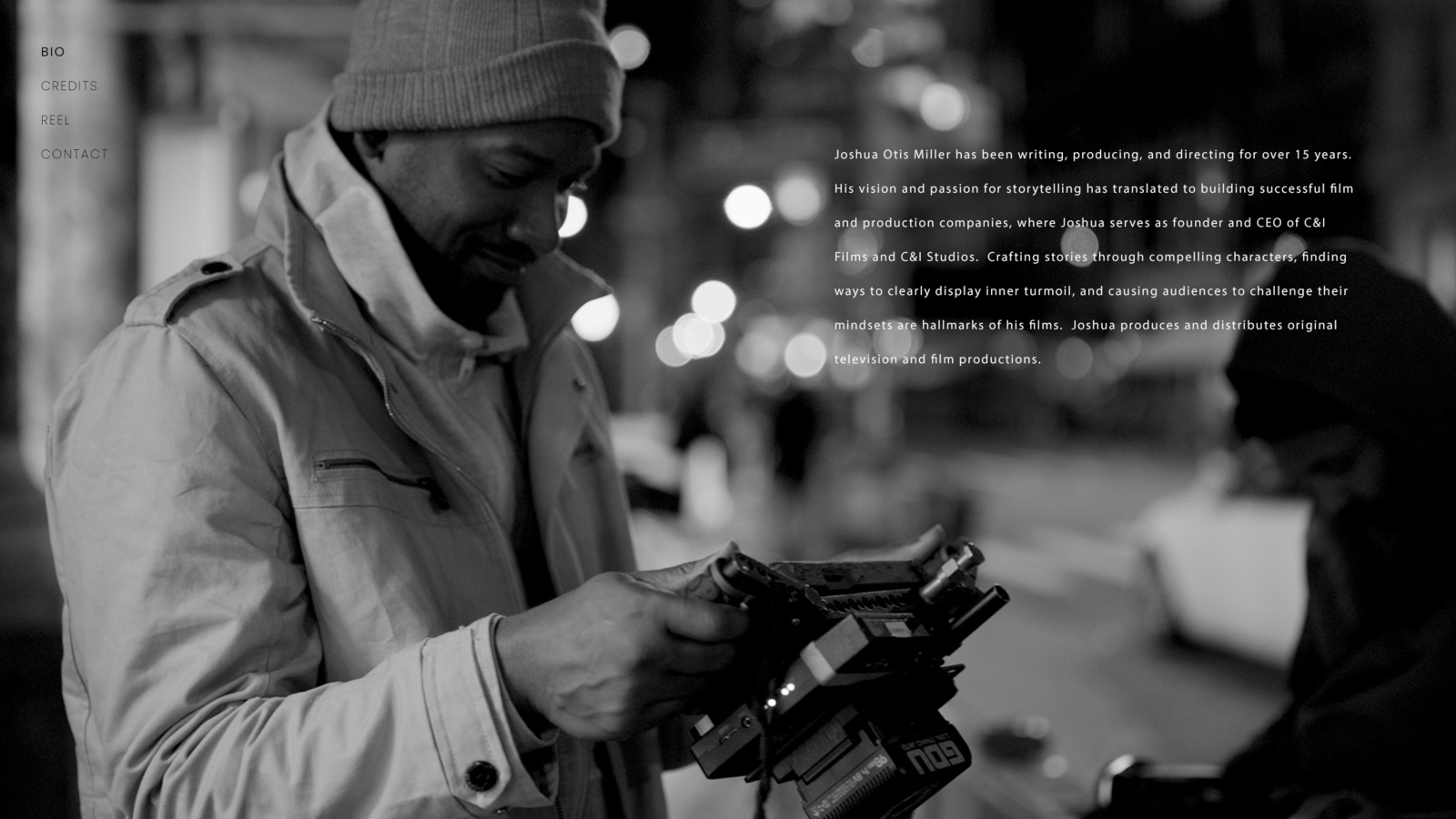 Black and white of Joshua wearing a knit cap and jacket looking at a camera video display smiling with white text
