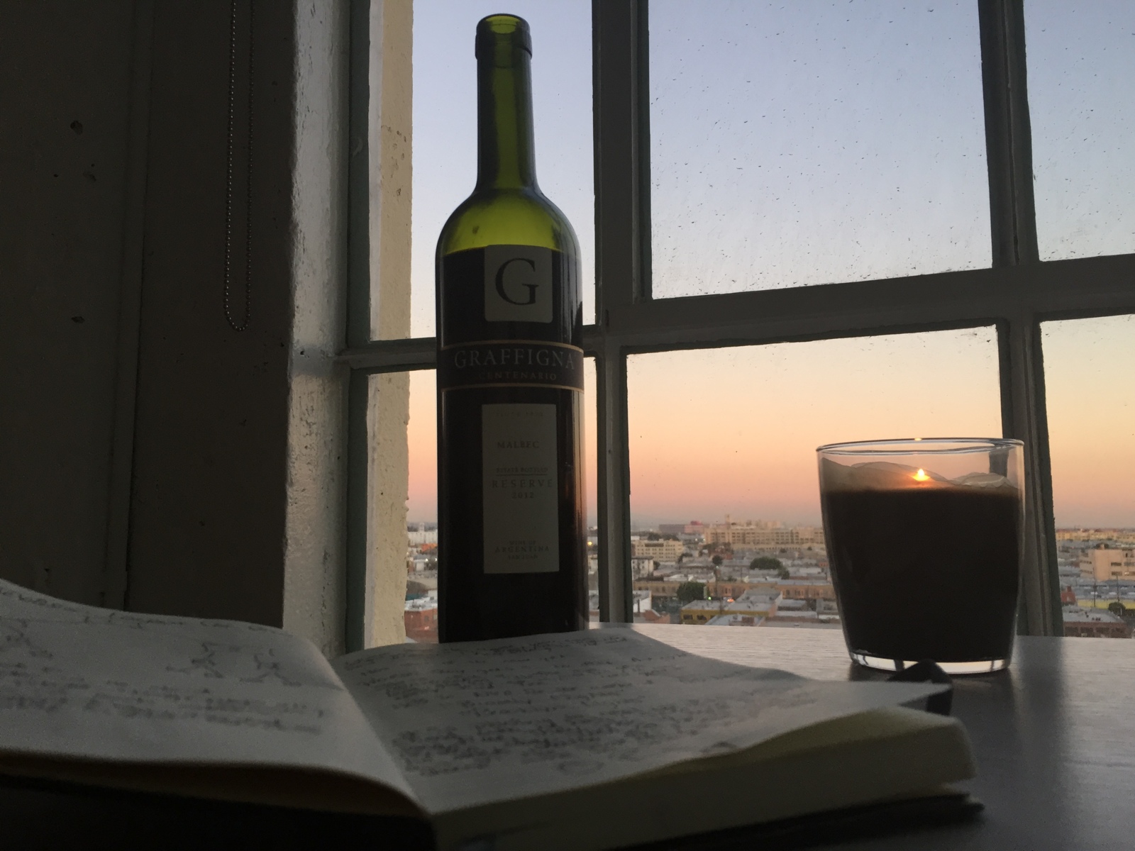 Bottle of Graffigna wine with opened notepad with notes and glass of coffee nearby looking out window at sun rising