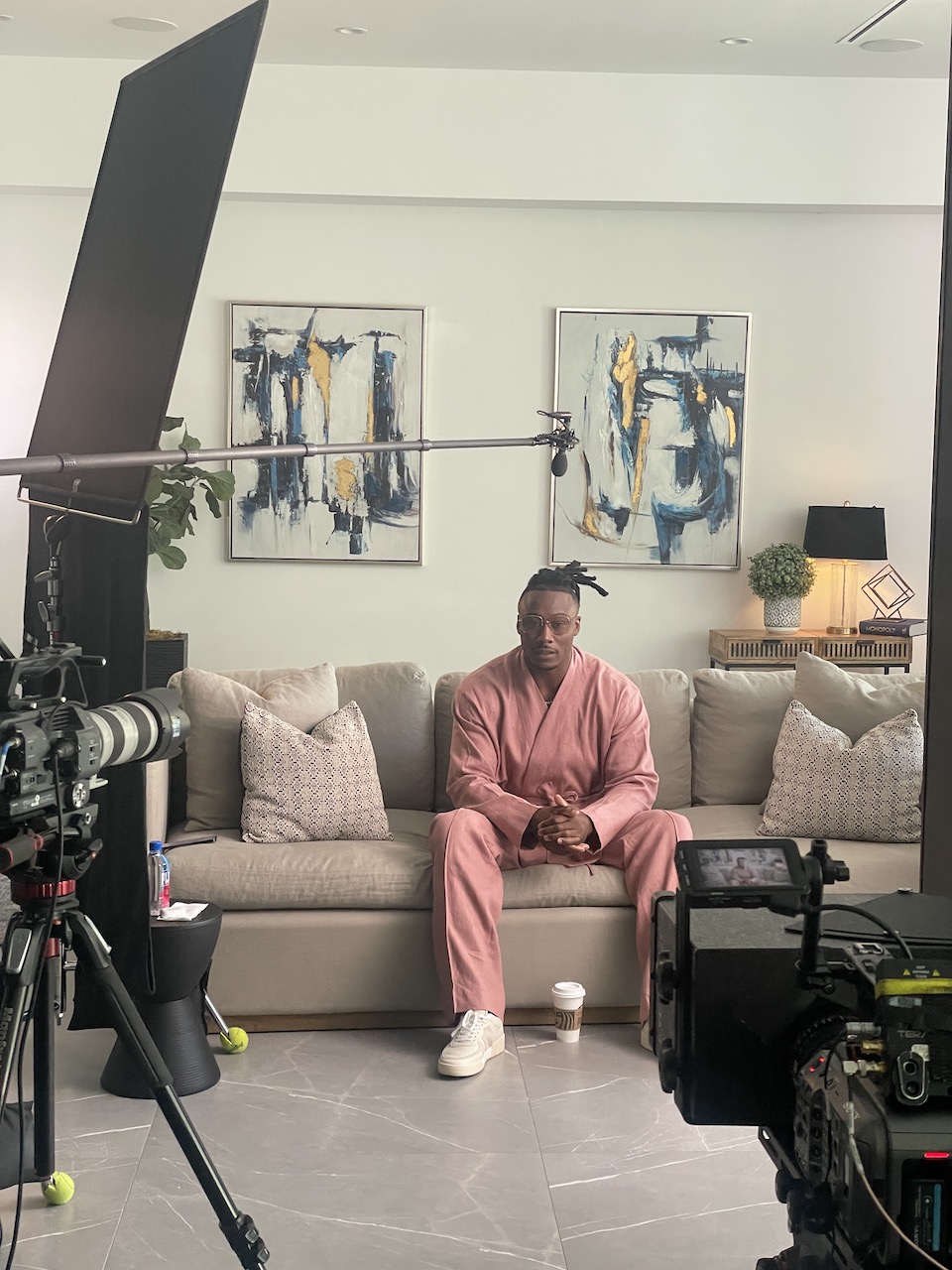 BlackPop BTS Brandon Marsall sitting on a brown couch posing for the camera wearing a pink outfit with two video cameras trained on him