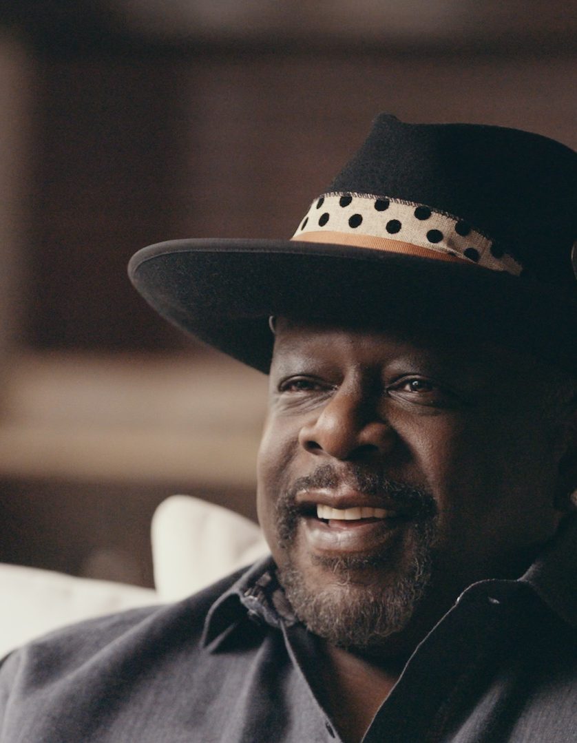 Black Pop Cedric The Entertainer Headshot Sitting On A White Couch Smiling Wearing A Black Hat And Gray Shirt