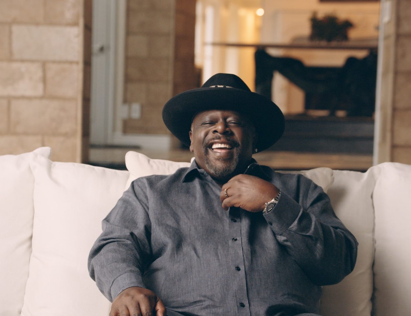 Black Pop Cedric the Entertainer headshot sitting on a white couch smiling wearing a black hat and gray shirt