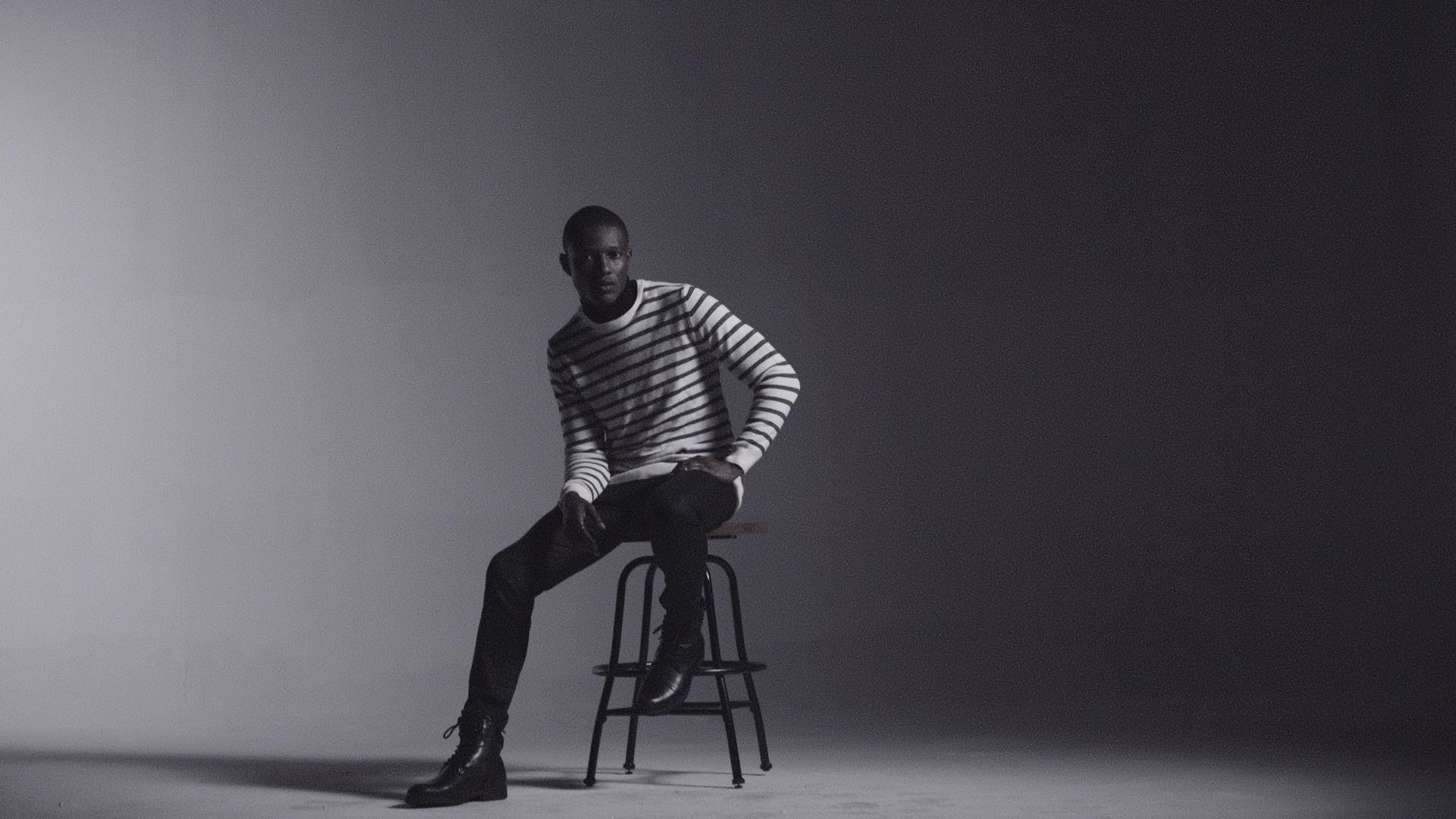 Black and white of African AMerican man sitting on a stool wearing a striped shirt with pants
