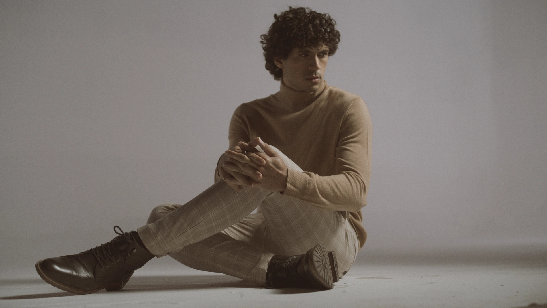 Man with curly hair wearing beige sweater and patterned pants