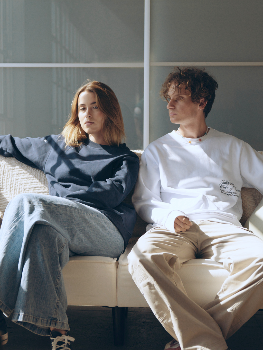 Female and male apparel models inside sitting on a couch wearing long sleeve shirts.